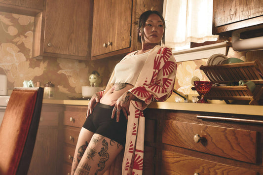 Confident woman in stylish black cotton boyshort underwear and cream top with bold red patterns, showcasing everyday comfort and edgy tattoo design in a vintage kitchen setting.