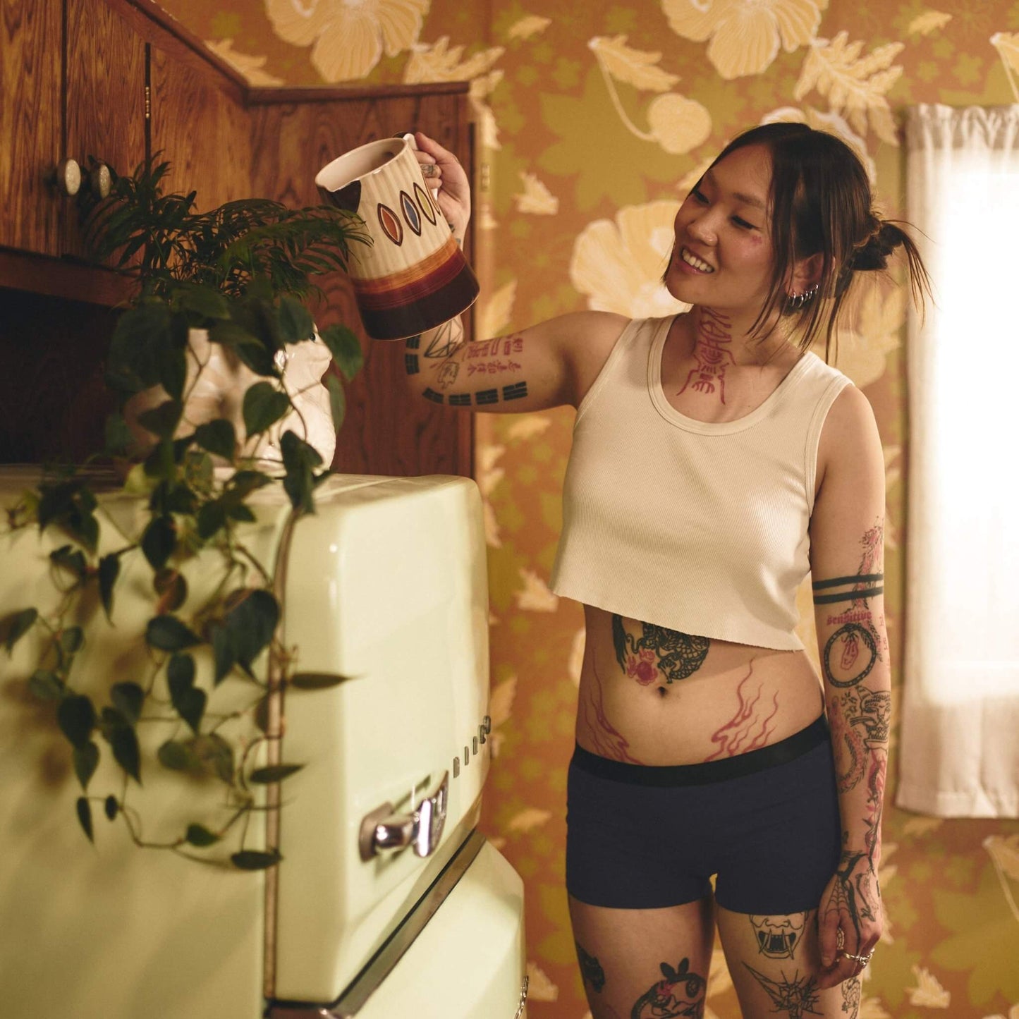 A woman with tattoos standing in front of a refrigerator wearing Better Clothing Company's Better Cotton Boyshort underwear.